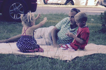 A father watching his children play while lying on a blanket in the grass waiting of a Fourth of July parade to start and for fireworks