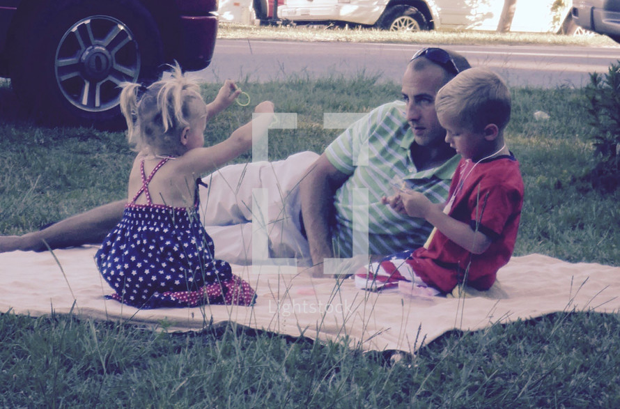 A father watching his children play while lying on a blanket in the grass waiting of a Fourth of July parade to start and for fireworks
