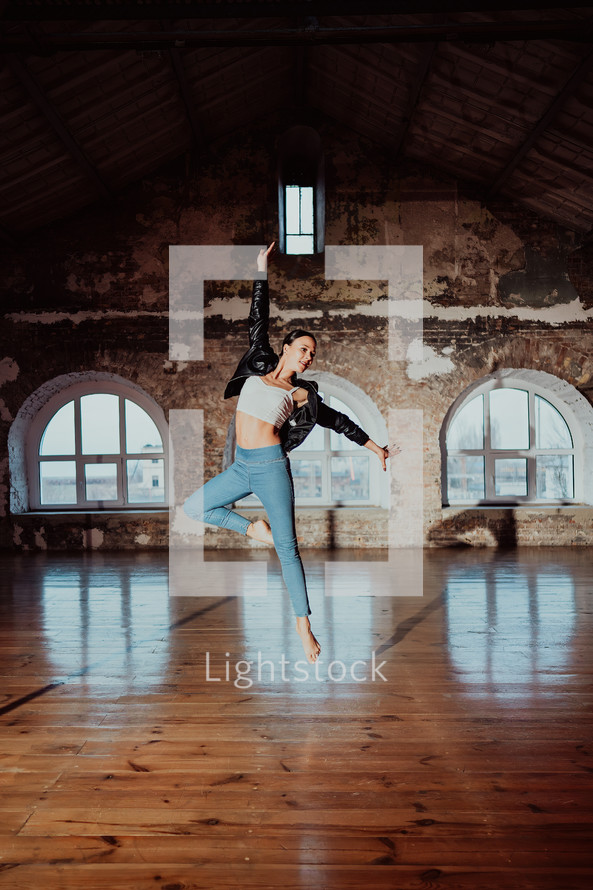Young woman jumping in casual style - jeans and leather jacket doing ballet in old studio. Attractive ballerina practices in choreography alone. High quality photo