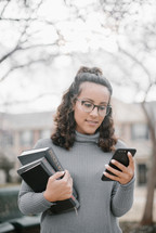 a young woman holding a stack of books and looking at her cellphone 
