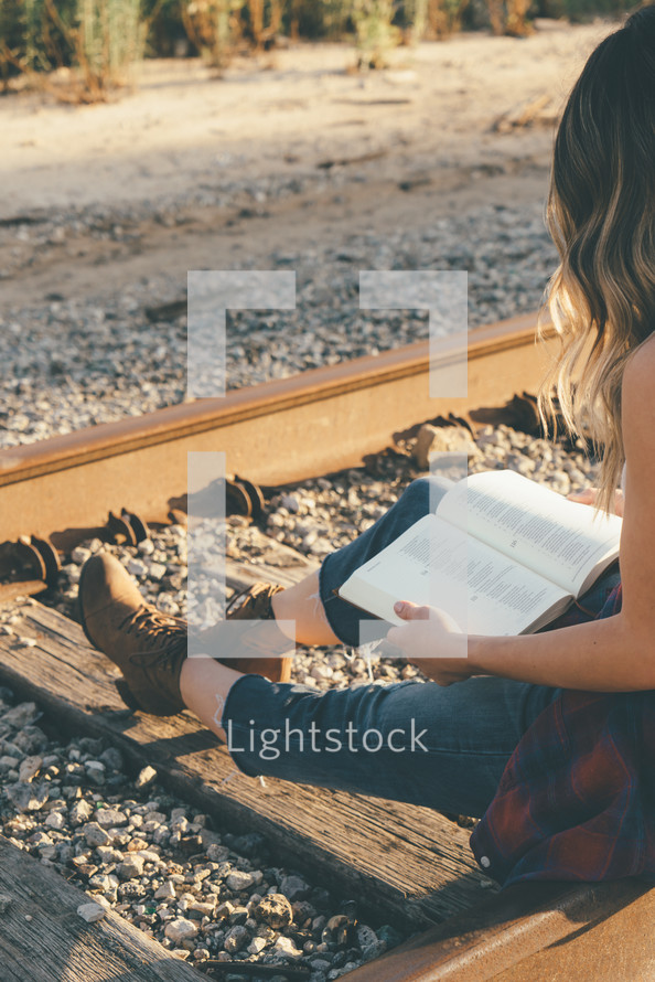 A young girl sitting on railroad tracks reading her bible.
