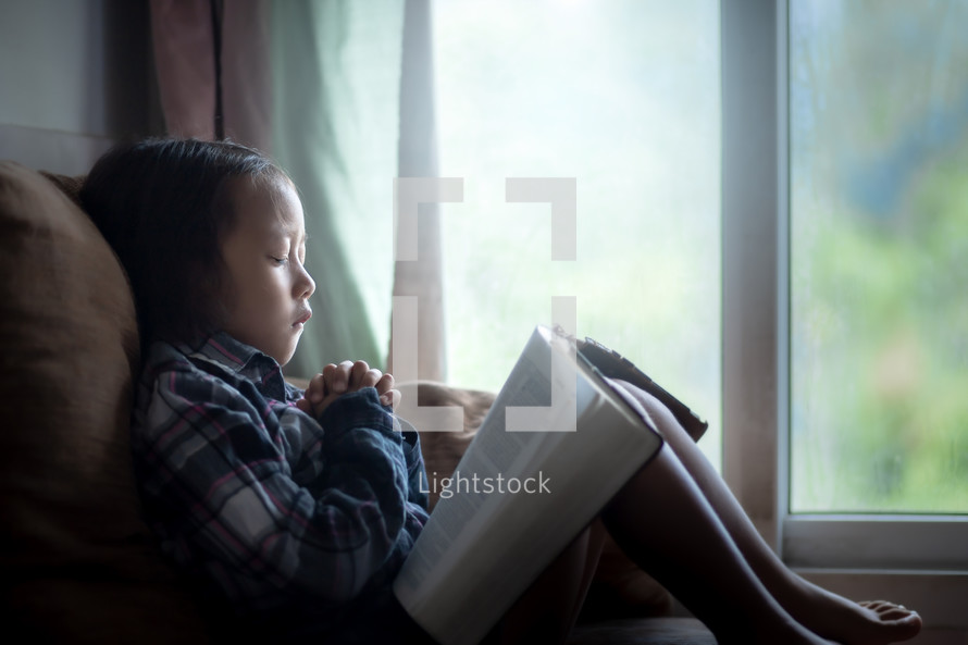 a little girl sitting in a chair reading a Bible and praying 