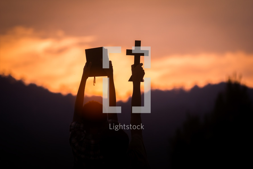 silhouette of a young man holding up a Bible and cross in worship at sunset 