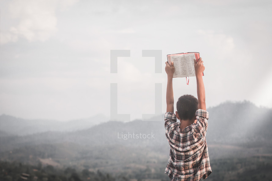 a boy holding up a Bible standing on a mountaintop 