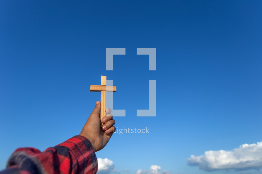 a arm holding up a wooden cross towards a blue sky 