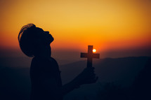 Young man praying and holding Cross at sunset  