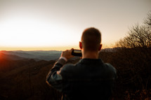 man taking a picture of mountains at sunrise 