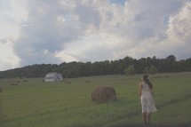 a woman in a dress and cowboy boots standing in a field with hay bales 