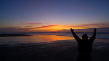 Man with his hands raised in praise and worship to God standing on a beach looking out at the ocean at sunset