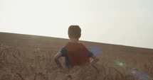 Young boy with a superhero cape stands in a golden field during sunset - raising hands in victory