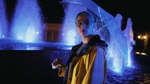 Young pretty girl with blue dyed hair in yellow raincoat and with transparent umbrella stands near fountain. Night illumination of city. Portrait of stylish hipster with glasses.
