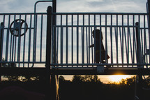 A young child (girl) runs across playground equipment with a sunset behind her.
