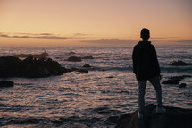 man standing on a rocky shore at sunset 
