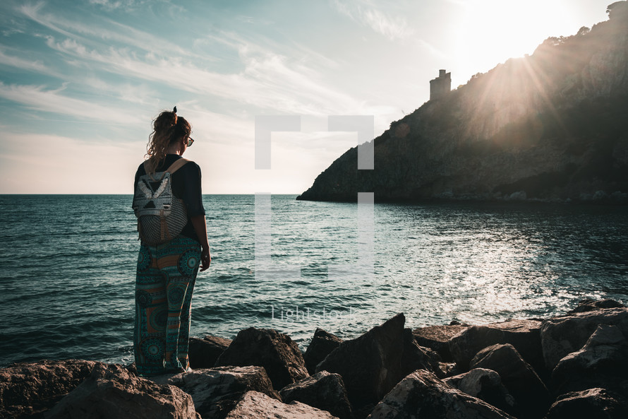 a girl with a backpack standing on a shore at sunrise 