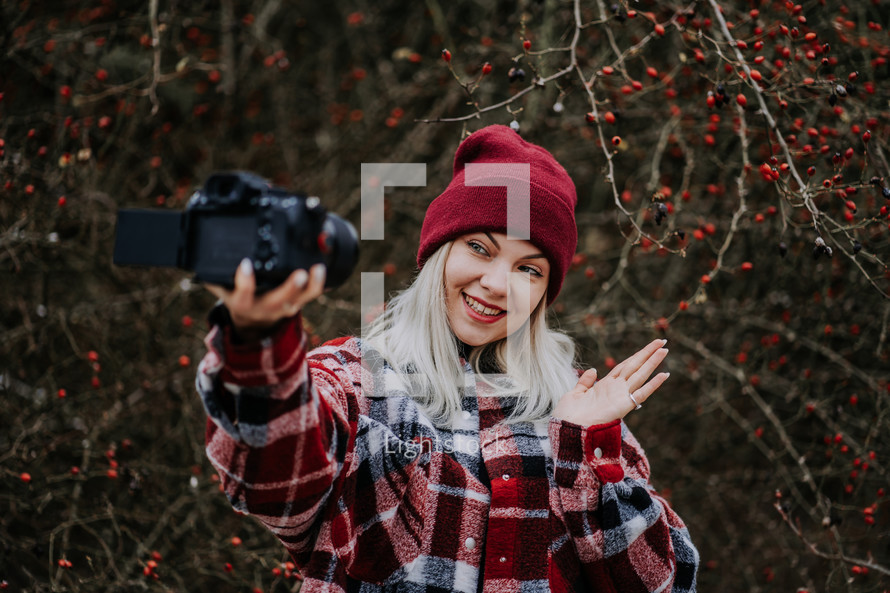 Portrait of young woman using camera in forest. Girl filming and smiling, lady in trendy shirt and red beanie hat. High quality