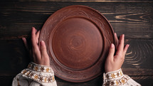 Female hands with empty ukrainian clay plate from wooden table. Woman in traditional embroidered shirt. Top view. National tableware with handmade ornament