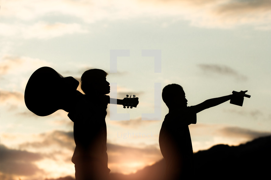 silhouettes of two boys holding a guitar and cross at sunset 