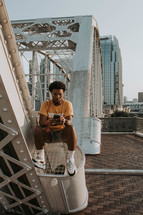 a man sitting on a bridge in a city texting 