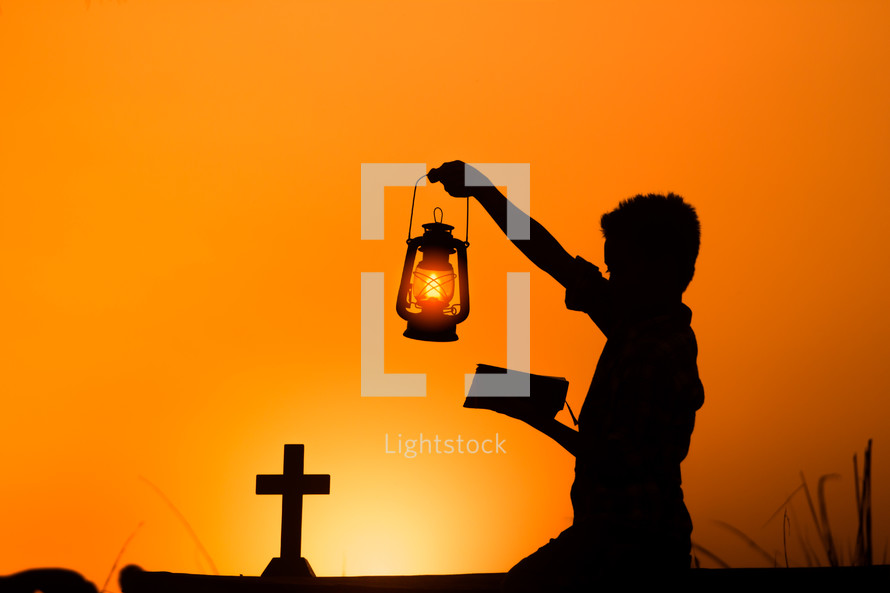 a silhouette of a boy holding a lantern and reading a Bible at sunset 