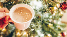hand holding hot chocolate in front of a Christmas tree 