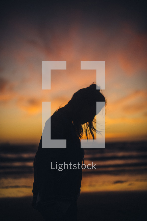 silhouette of a woman looking down standing on a beach at sunset 
