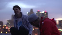 young men sitting on a roof talking above a city 