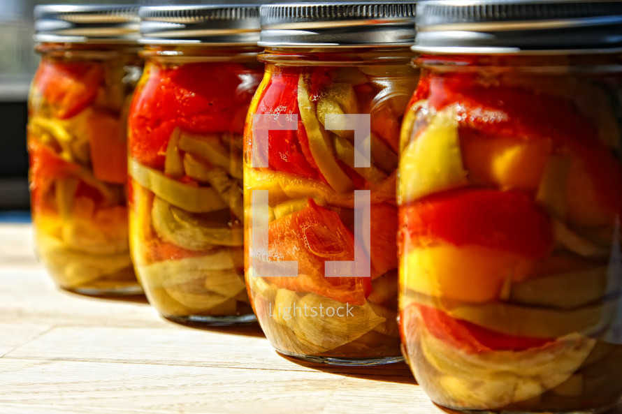 Jars of home canned organic spicy and sweet peppers sitting on the counter top in sunlight.  Healthy and nutritional food.
  (Shallow depth of field, focus point on second jar from right).