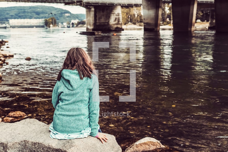 a girl sitting on a rock by a river 