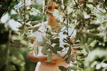 a woman standing behind green leaves on a tree branch hidden 