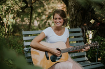 a woman sitting on a bench playing an acoustic guitar 