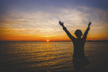 silhouette of a woman with arms raised in worship in front of the ocean 