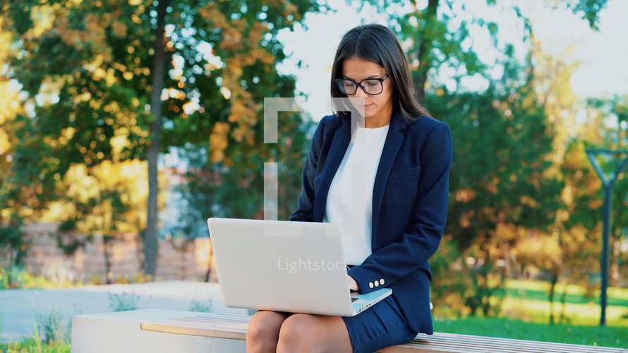 Successful business woman with laptop computer on green park background. Beautiful girl in formal wear and glasses surfing internet or working on electronic device
