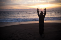 A woman raising her hands on the beach at sunset.