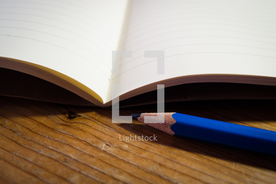 An open blank notebook with a blue pencil laying next to it on a wooden desk