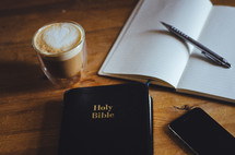 A coffee, phone, notebook, pen and bible on a table