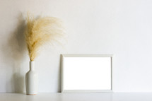 vase of brown fuzzy grasses and blank frame 