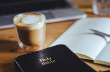 A coffee, bible, notebook and laptop on a table.