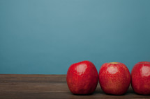 Three red apples lined up on a wooden surface with a blue background.