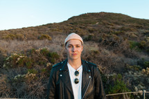 man in a beanie and leather jacket standing on a hill 