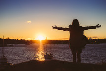 Silhouette of a woman with arms extended at sunset on a lake.