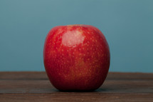 A lone red apple on a wooden surface with a blue background.