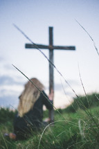 Woman kneeling in worship at a wooden cross in a field of grass.