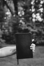 Tattooed arm holding a Bible outside.