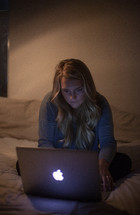 young woman sitting on her bed looking at a computer screen in a dark room 