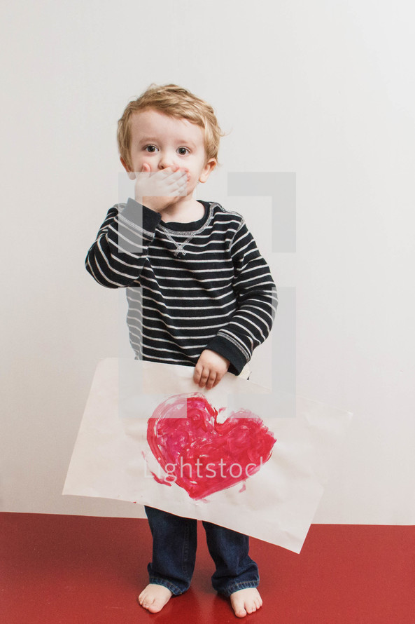boy child with his hands over his mouth holding a Valentines picture 
