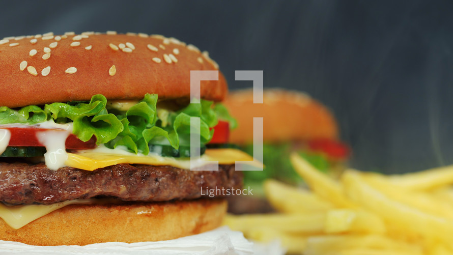 Big appetizing burger with meat cutlet, vegetables, cheese, lettuce and sauce. Hamburger rotates on other meal background, close-up view. Unhealthy yummy food concept.