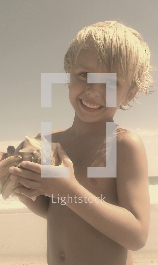 A happy little boy holding a whelk seashell on a beach on a summer day 