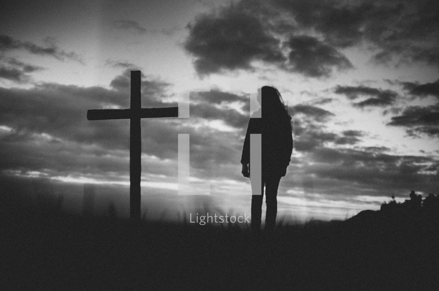 Silhouette of a person standing by a cross in a field at sunrise.