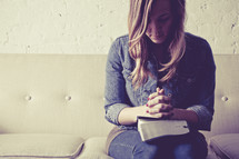 woman sitting on a couch with her hands in prayer held over a Bible.