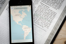 A phone with a world map showing North and South America sitting on a bible open to the great commission